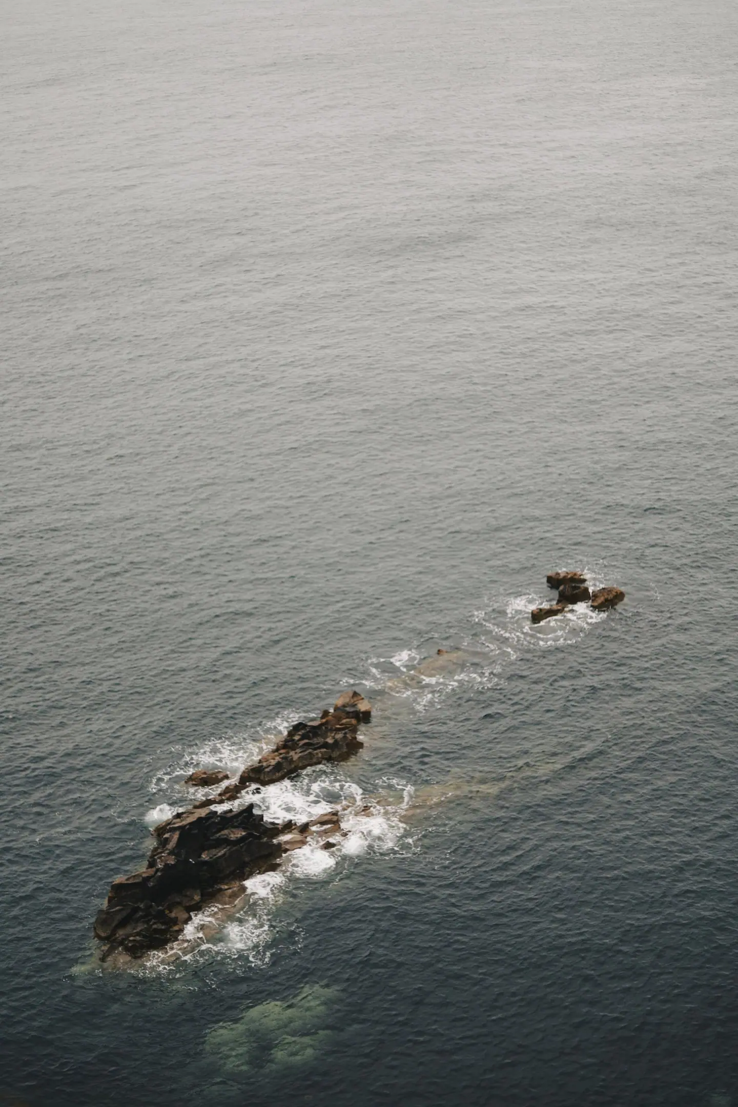 A line of dark rocks and white surf in a calm grey sea off the Scottish coast.